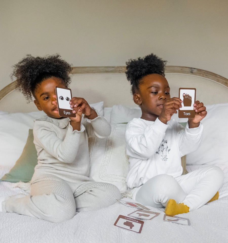 Two young children sitting on a bed holding up Parts of the Body flashcards